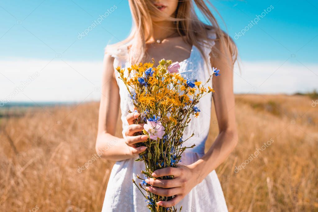 cropped view of young woman in white dress holding wildflowers in grassy field, selective focus