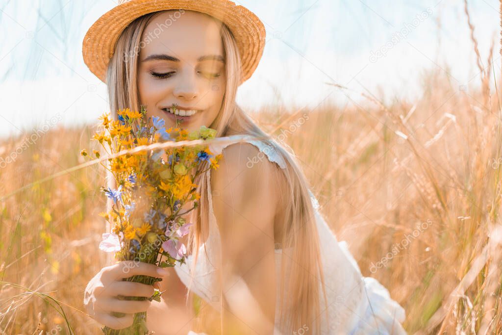 selective focus of sensual woman in straw hat holding wildflowers in grassy field