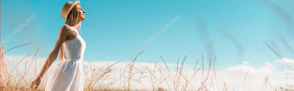 selective focus of young woman in white dress and straw hat standing with outstretched hands against blue sky, panoramic shot