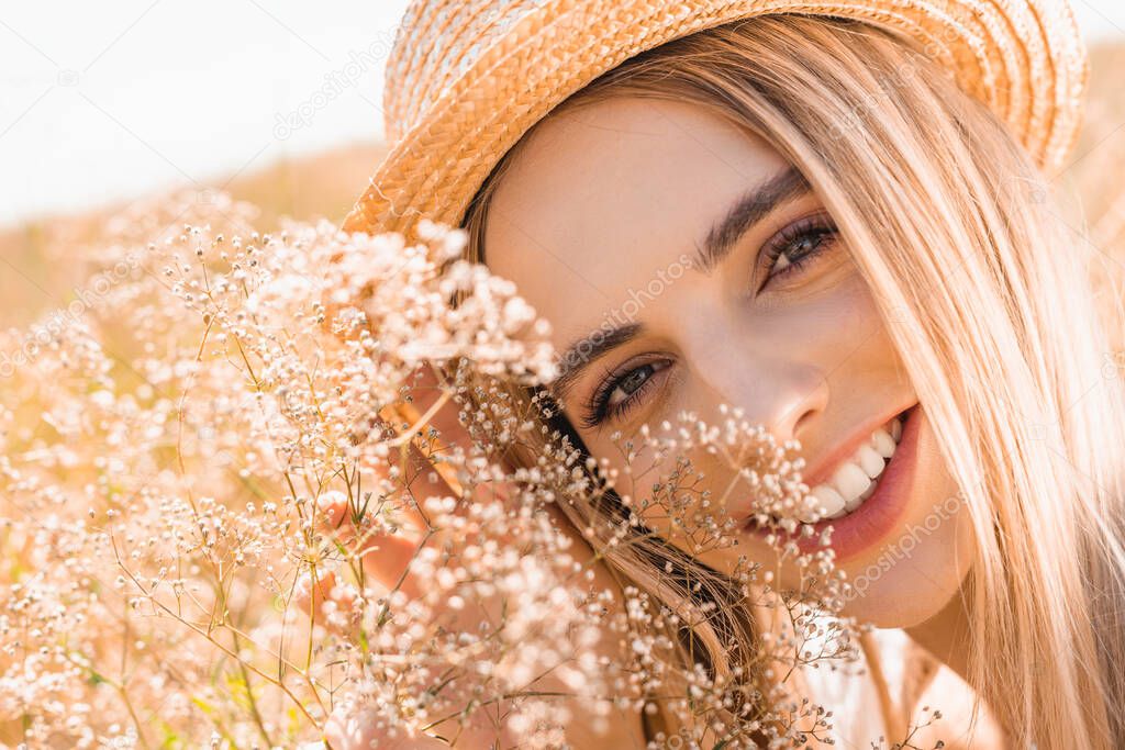 portrait of young blonde woman in straw hat looking at camera near wildflowers, selective focus