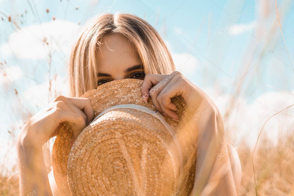 selective focus of blonde woman obscuring face with straw hat while looking at camera against blue sky