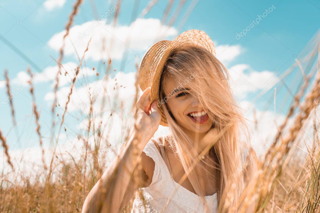 selective focus of excited blonde woman touching straw hat and looking at camera against blue cloudy sky