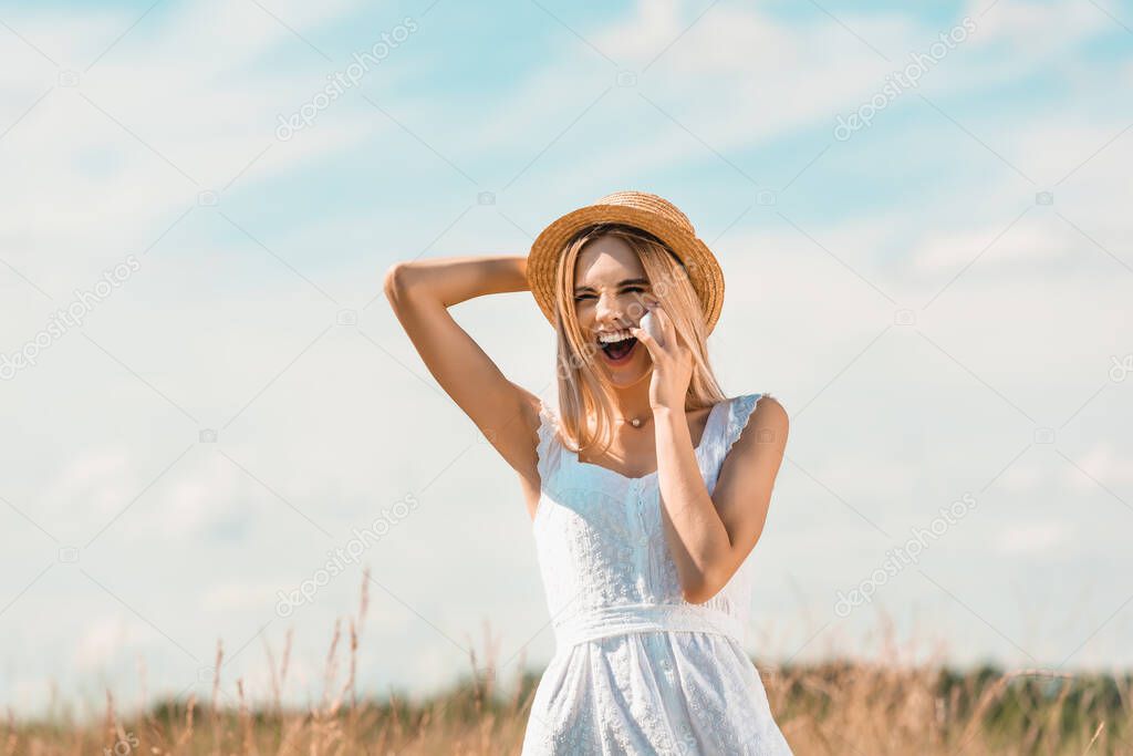 excited blonde woman in white dress touching straw hat and laughing in field against blue sky