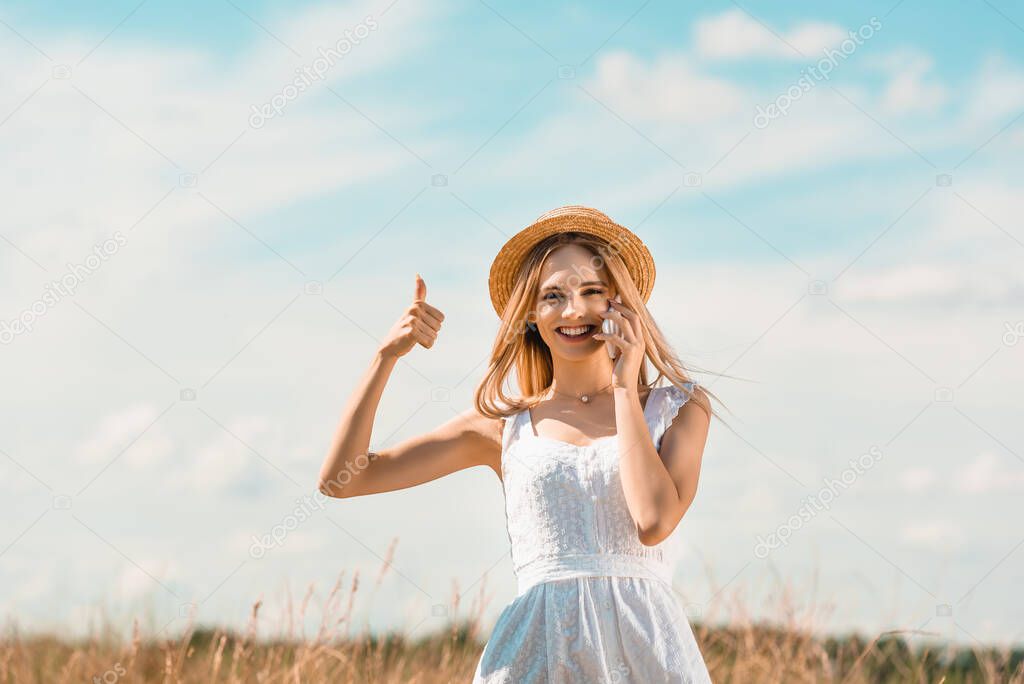 excited woman in white dress and straw hat showing thumb up while looking at camera against blue sky