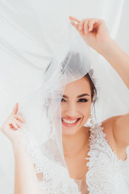 Selective focus of bride in wedding dress holding veil and looking at camera near white cloth  clipart