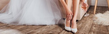 Panoramic shot of bride in wedding shoes holding glass of wine near dress at home  clipart