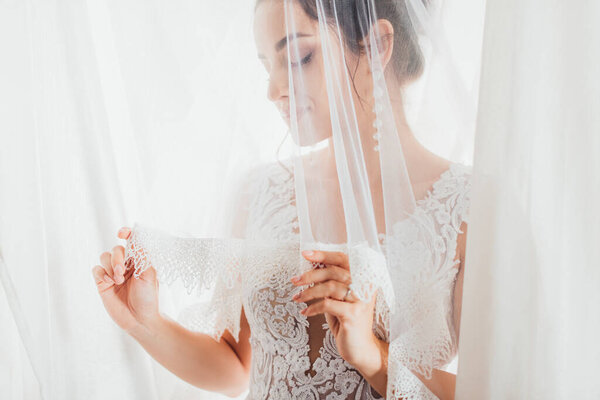 Selective focus of young bride holding lace veil near white curtains