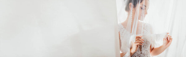 Panoramic shot of bride touching veil near white curtains 