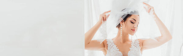 Panoramic crop of bride in wedding dress and pearl earrings holding veil near white cloth 