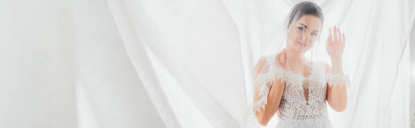 Horizontal crop of bride in wedding dress and veil looking at camera near white curtains 