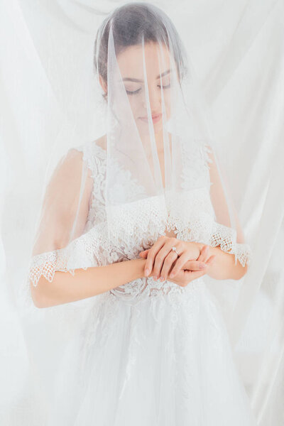 Young bride in lace wedding dress and veil standing near white cloth 