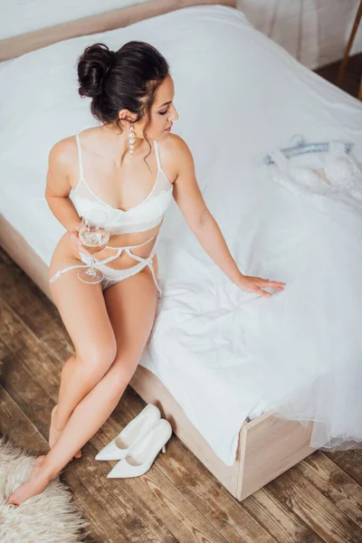 ランジェリーで花嫁のオーバーヘッドビューワインのグラスを保持し ベッドの上で結婚式のドレスに触れる — ストック写真