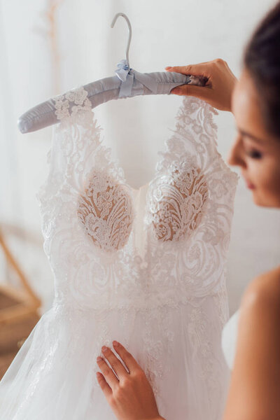 Selective focus of bride holding lace wedding dress on hanger at home 