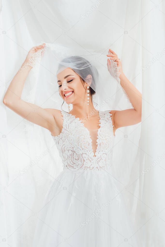 Brunette bride in lace wedding dress and pearl earrings holding veil near white cloth 