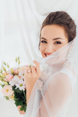 Perdelerin yanındaki kameraya bakarken duvak ve buket tutan genç gelin. 