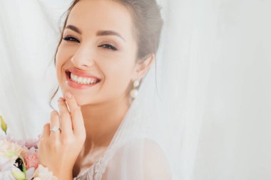 Genç gelinin seçici odağı buket ve perdelerin yanındaki kameraya bakıyor. 