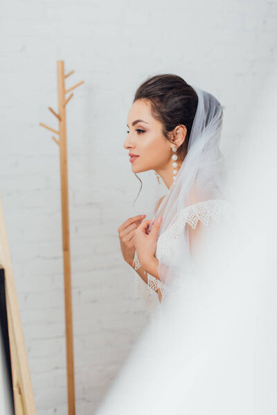 Selective focus of brunette bride in pearl earring wearing veil at home 