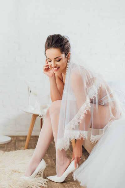 Brunette bride in underwear and veil wearing heels beside wedding dress in bedroom