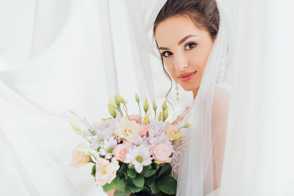 Selective focus of bride in veil holding floral bouquet and looking at camera near curtains 