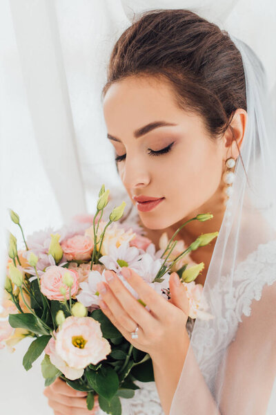 Selective focus of young bride in wedding dress and veil touching flowers beside curtains 