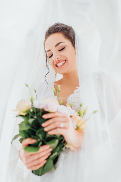 Selective focus of young bride in veil and dress holding floral bouquet 