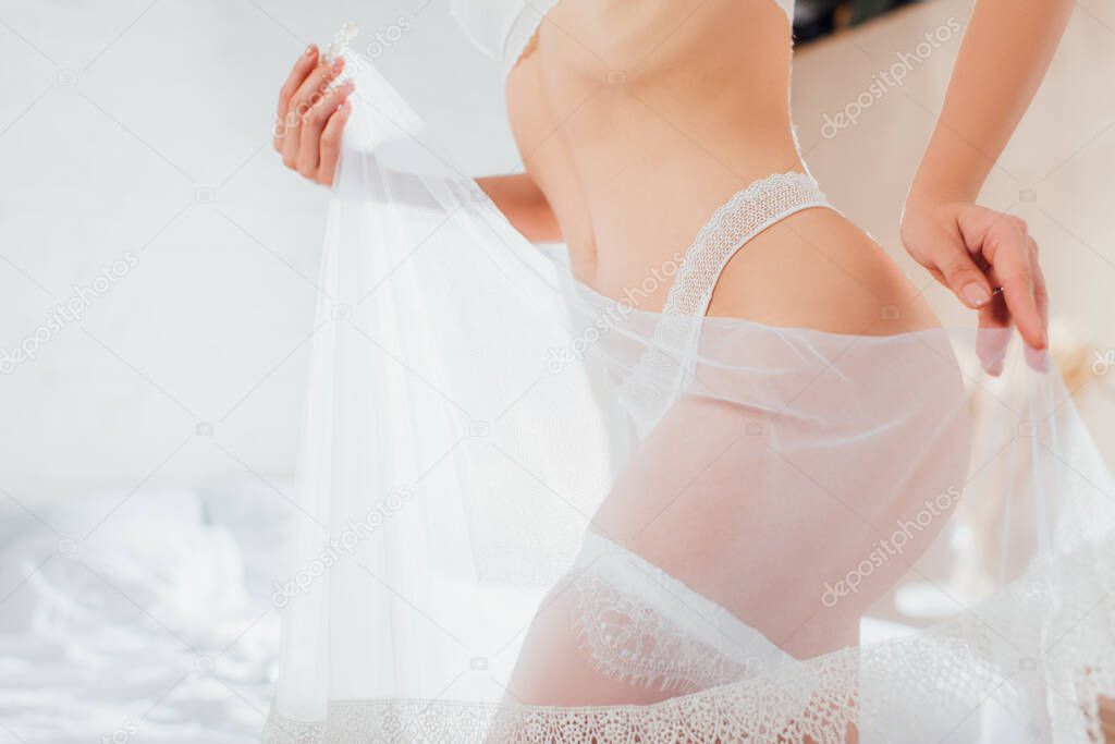 Cropped view of sensual bride in underwear and garter holding veil in bedroom