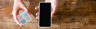 Panoramik kadın elinde boş ekranlı akıllı telefon ve ilaç organizatörüyle ilaç tutuyor.