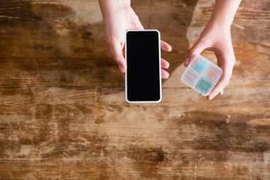 Elinde boş ekranlı akıllı telefon ve ilaç organizatörü olan bir kadının üst görüntüsü.