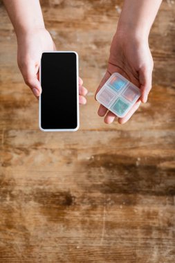 Genç bir kadının elinde boş ekranlı akıllı telefon ve ilaç organizatörüyle ilaç tutuşu.