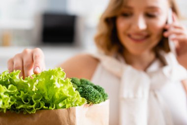 selective focus of woman touching fresh lettuce near broccoli while talking on smartphone  clipart