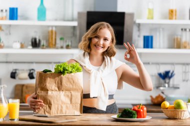 young woman showing ok sign near paper bag with lettuce and bowl with fruits clipart