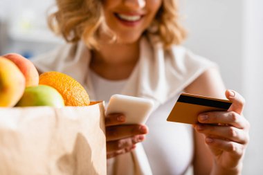 Akıllı telefon ve kredi kartını kağıt torbanın içinde meyvelerin yanında tutan kadının kısmi görüntüsü 