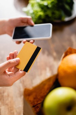 Seçici odak noktası kredi kartı ve akıllı telefon elinde boş ekranlı kağıt torba içindeki meyvelerin yanında 