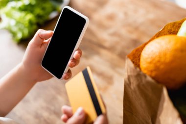 Seçici odak noktası, elinde boş ekranlı akıllı telefon ve kağıt torbada meyvelerin yanında kredi kartı olan kadın. 