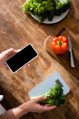 Mutfak pullarının yanında brokoli tutan bir kadın ve boş ekranlı akıllı telefon. 