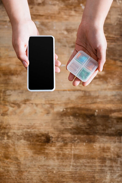 top view of young woman holding smartphone with blank screen and medication in pill organizer
