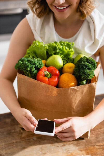 cropped view of woman holding smartphone with blank screen near groceries 