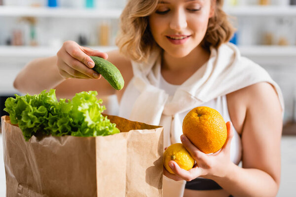 selective focus of woman holding cucumber, lemon and orange near paper bag 