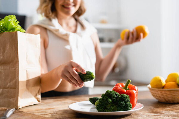 cropped view of young woman holding cucumber near plate with broccoli and bell pepper