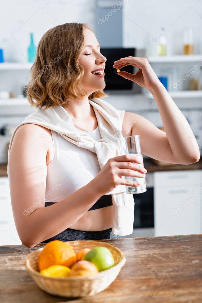 young woman taking pill and holding glass of water near fruits 