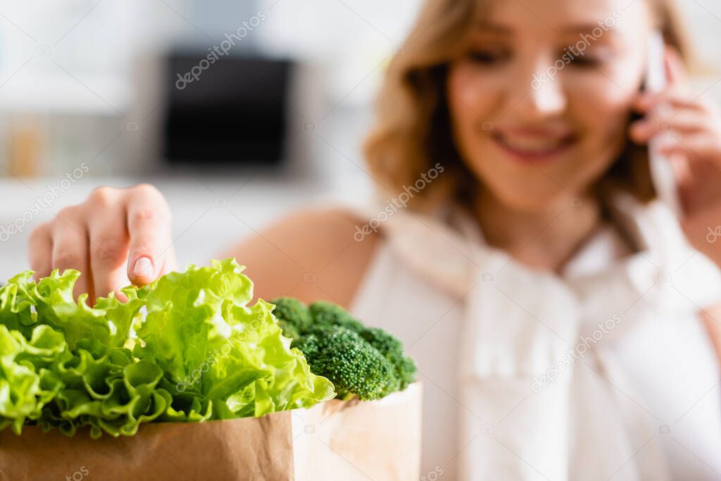 selective focus of woman touching fresh lettuce near broccoli while talking on smartphone 