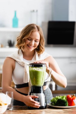 Sebzelerin yanındaki karıştırıcıda yeşil smoothie karıştıran genç bir kadın. 