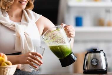 Kırık manzaralı genç bir kadın mikserle cam kenarında yeşil smoothie tutuyor. 