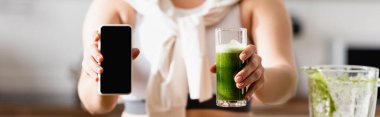 Elinde bir bardak yeşil smoothie ve boş ekranlı akıllı telefondan oluşan panoramik ekin.
