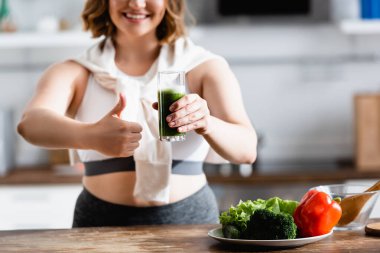 Genç bir kadının elinde bir bardak yeşil smoothie tutarken baş parmağını göstermesi. 