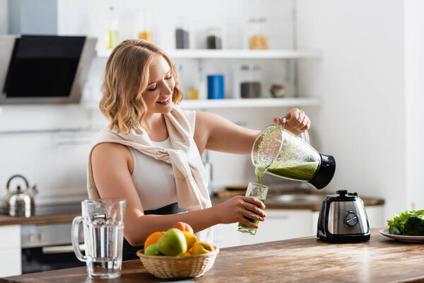 selective focus of woman pouring green smoothie in glass near fruits
