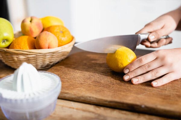 cropped view of woman cutting lemon on chopping board near fruits and juicer