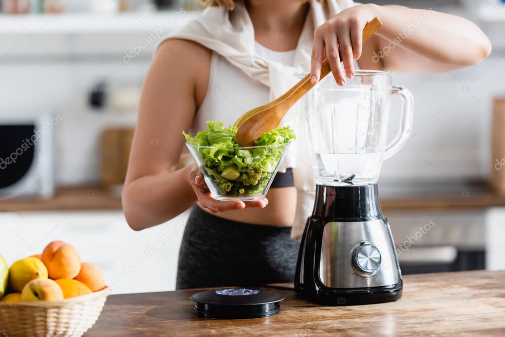 cropped view of woman holding bowl with lettuce and wooden spoon near blender 
