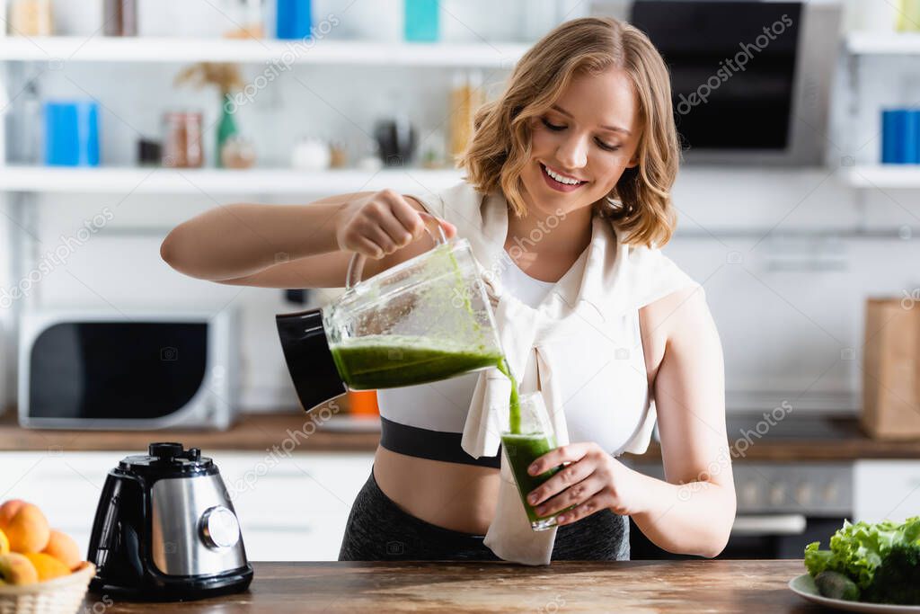 selective focus of young woman pouring green smoothie in glass near fruits