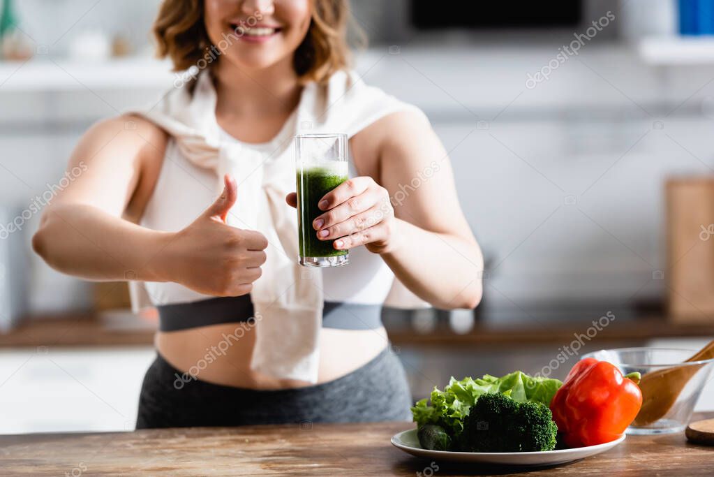 cropped view of young woman holding glass of green smoothie while showing thumb up 